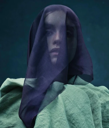 Portrait of a person with a purple sheer veil over their face, wearing a green textured garment against a dark background.