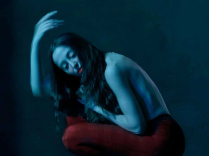 Dramatic studio portrait of a woman in a crouched pose, illuminated with blue and red lighting against a dark background.