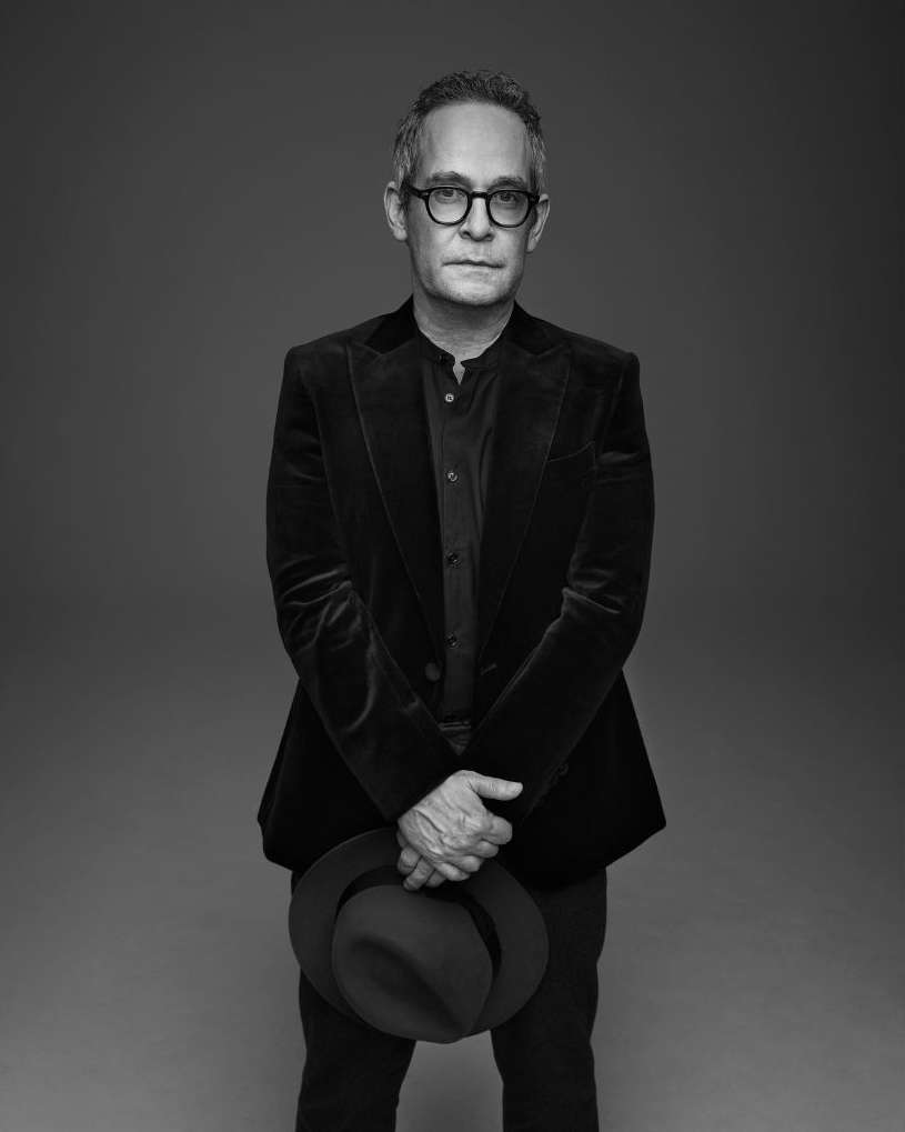 Black and white portrait of a mature adult man wearing glasses and a dark blazer, holding a dark hat in front of a grey backdrop.