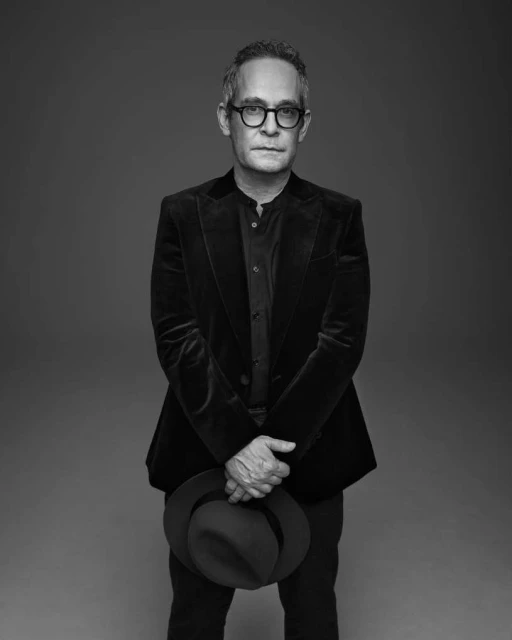 Black and white portrait of a mature adult man wearing glasses and a dark blazer, holding a dark hat in front of a grey backdrop.