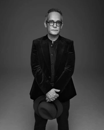 Black and white portrait of a mature adult man wearing glasses and a dark blazer, holding a dark hat in front of a grey backdrop.