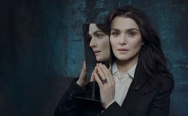 Rachel Weisz holding a rectangular mirror reflecting her profile against a dark textured background