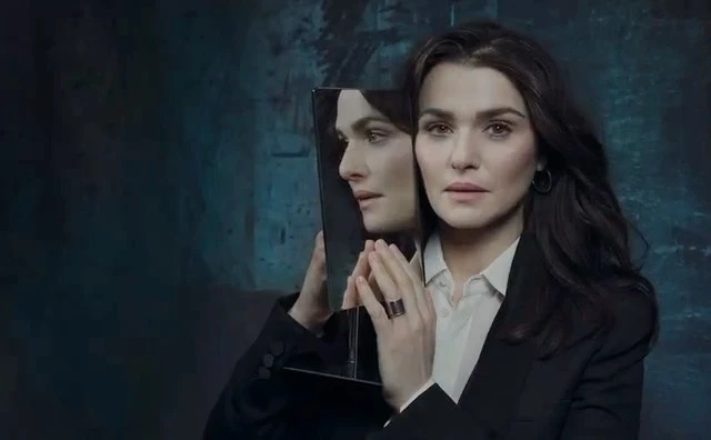 Rachel Weisz holding a rectangular mirror reflecting her profile against a dark textured background