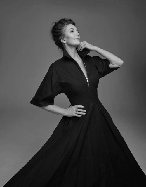 Portrait of a woman in a dark dress with a zipper, collar, and flared skirt. She has her hand on her hip and is posing indoors.