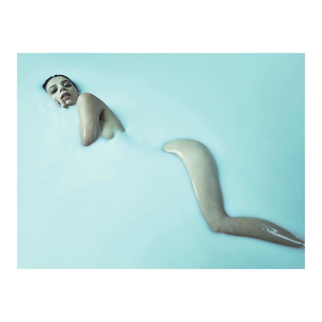 Artistic image of a woman partially submerged in milky blue water, focusing on her head, shoulders, arms, and partially visible leg.
