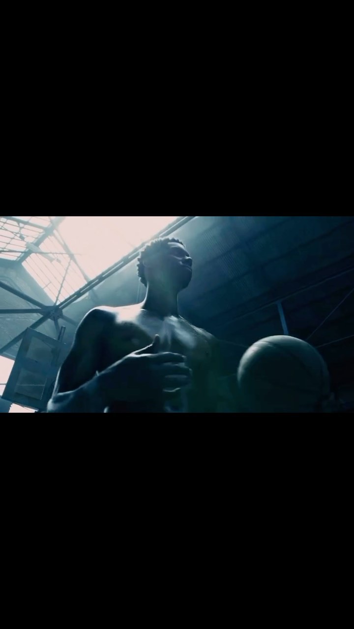African American man holds basketball in a dark, industrial setting with skylight overhead. Low angle captures upward gaze, athletic physique.