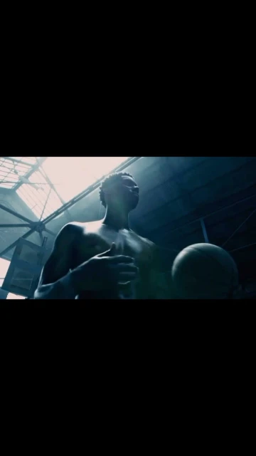 African American man holds basketball in a dark, industrial setting with skylight overhead. Low angle captures upward gaze, athletic physique.