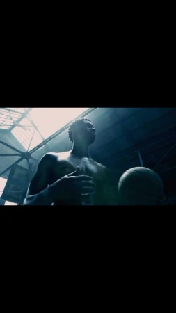 African American man holds basketball in a dark, industrial setting with skylight overhead. Low angle captures upward gaze, athletic physique.