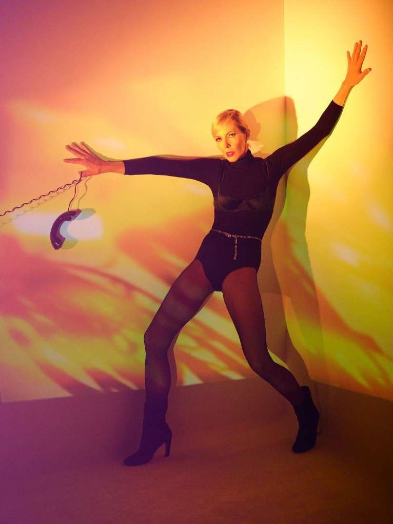 Woman strikes a pose in a black bodysuit with a telephone in an indoor studio, bathed in vibrant yellow and orange lighting.