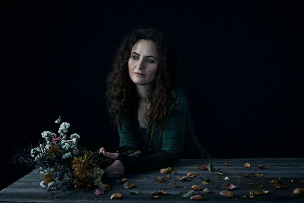 Portrait of a woman with long curly hair seated at a wooden table with a bouquet of dried flowers and scattered petals.