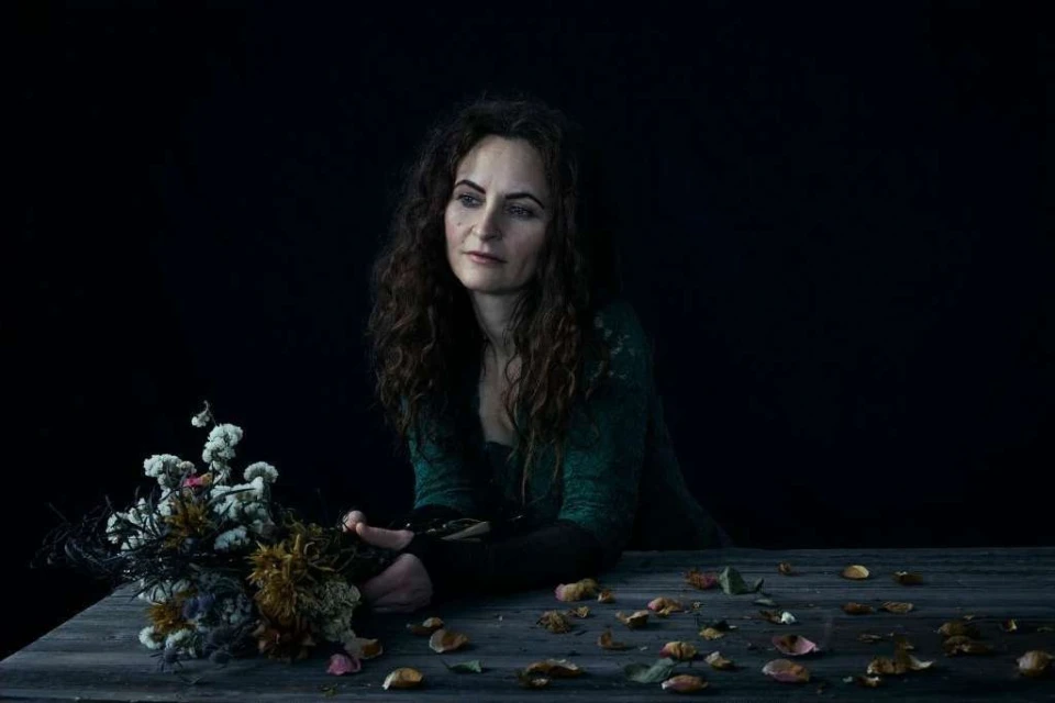 Portrait of a woman with long curly hair seated at a wooden table with a bouquet of dried flowers and scattered petals.