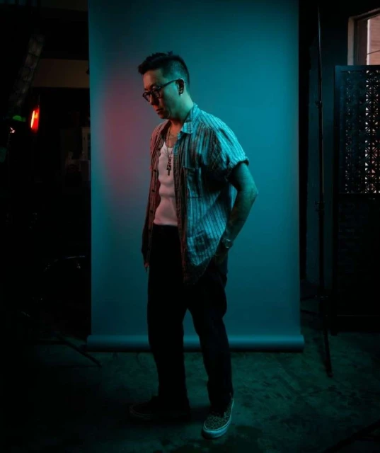 Adult man in glasses, striped shirt, and dark pants poses against a cyan backdrop in a studio with minimal lighting.
