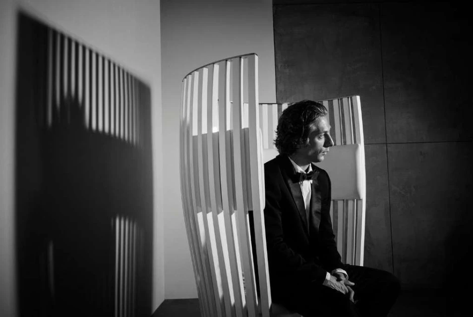Elegant man in a black tuxedo seated in a sculptural chair, illuminated by soft studio lighting, casting shadow patterns.