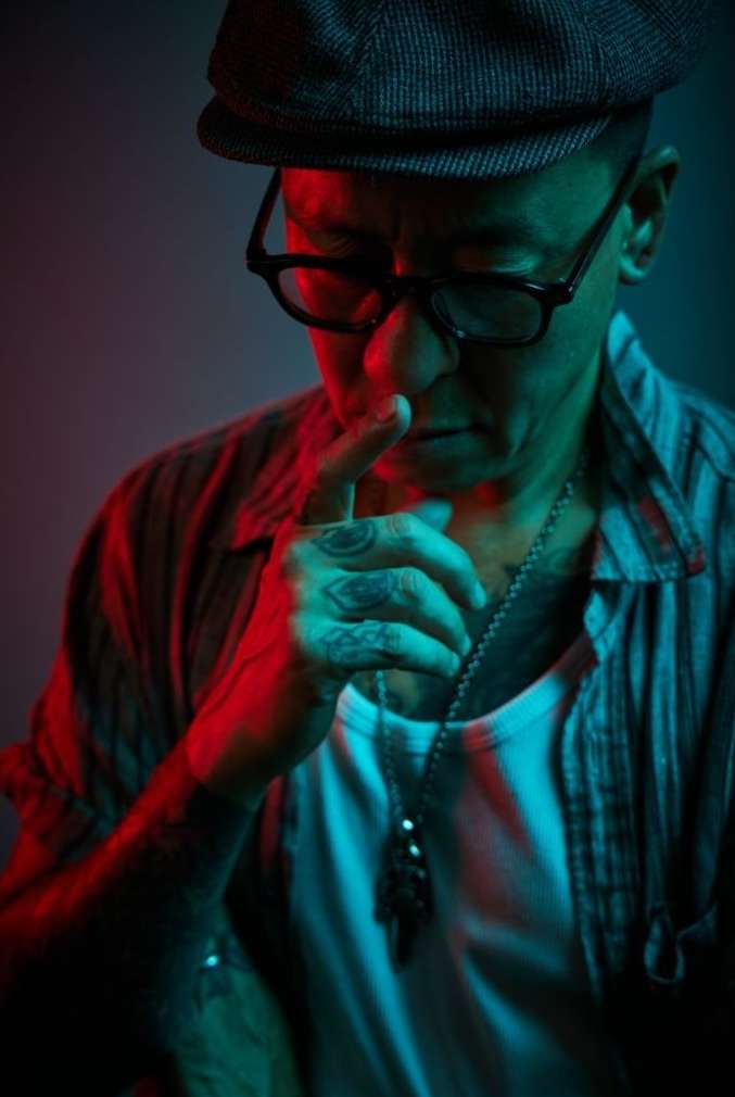 Man wearing flat cap, glasses, and layered shirts with finger touching his nose in dramatic red and teal lighting.