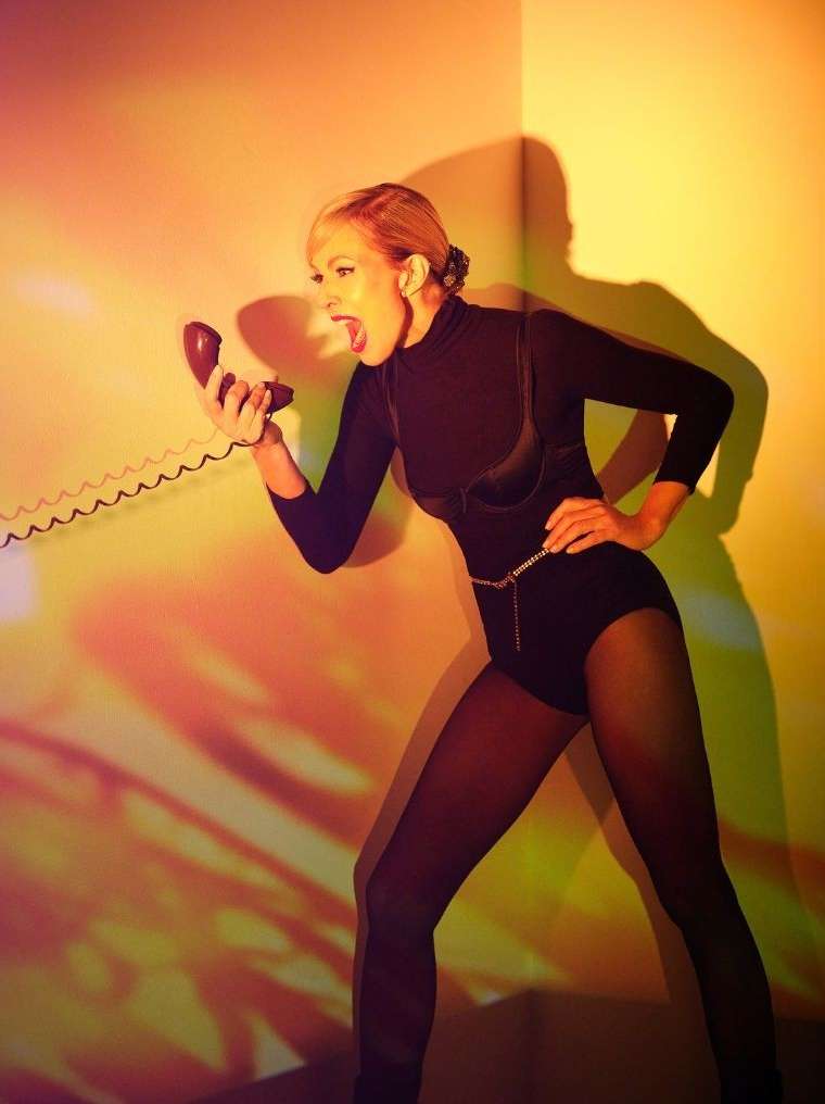 A woman in a black bodysuit and tights shouts into a red rotary phone in a dramatic pose in front of an orange and yellow wall.