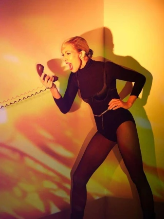 A woman in a black bodysuit and tights shouts into a red rotary phone in a dramatic pose in front of an orange and yellow wall.