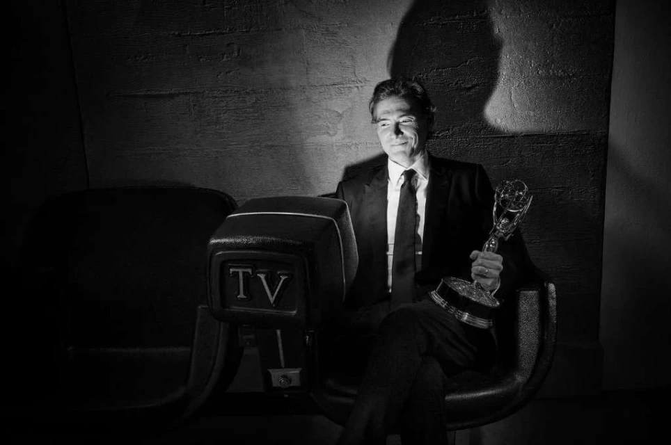 Black and white shot of mature man in suit sitting indoors, holding award with vintage TV set element in dark, textured environment.