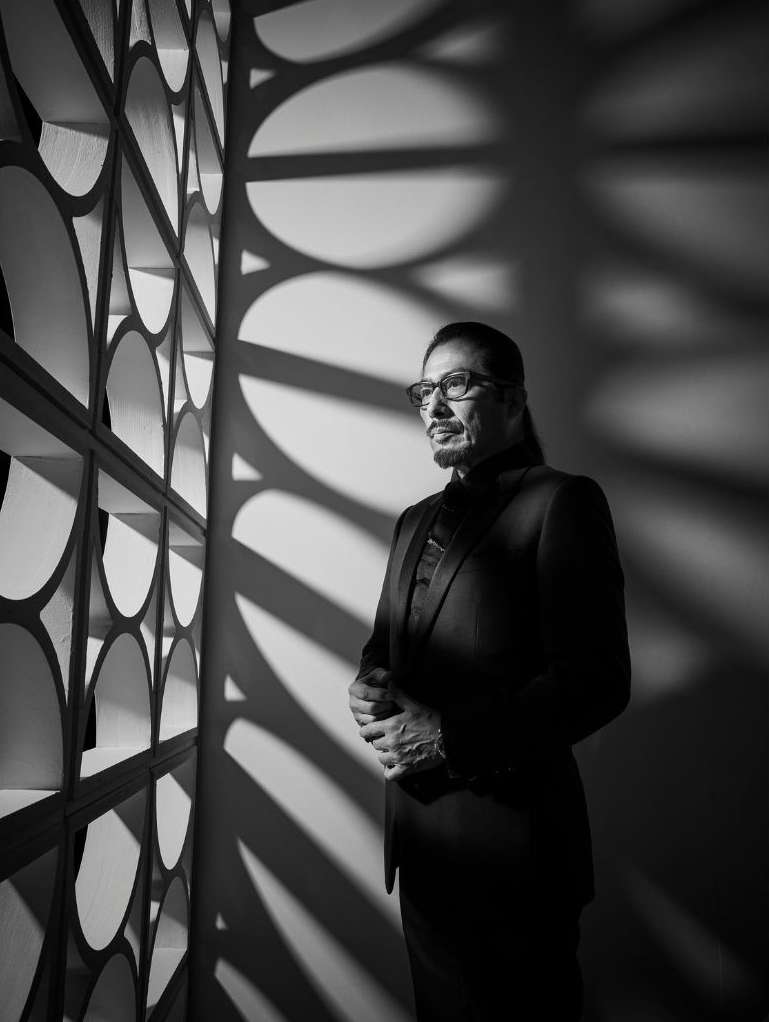 Black and white portrait of an adult male in a suit and glasses, standing indoors next to a patterned wall, with shadows.