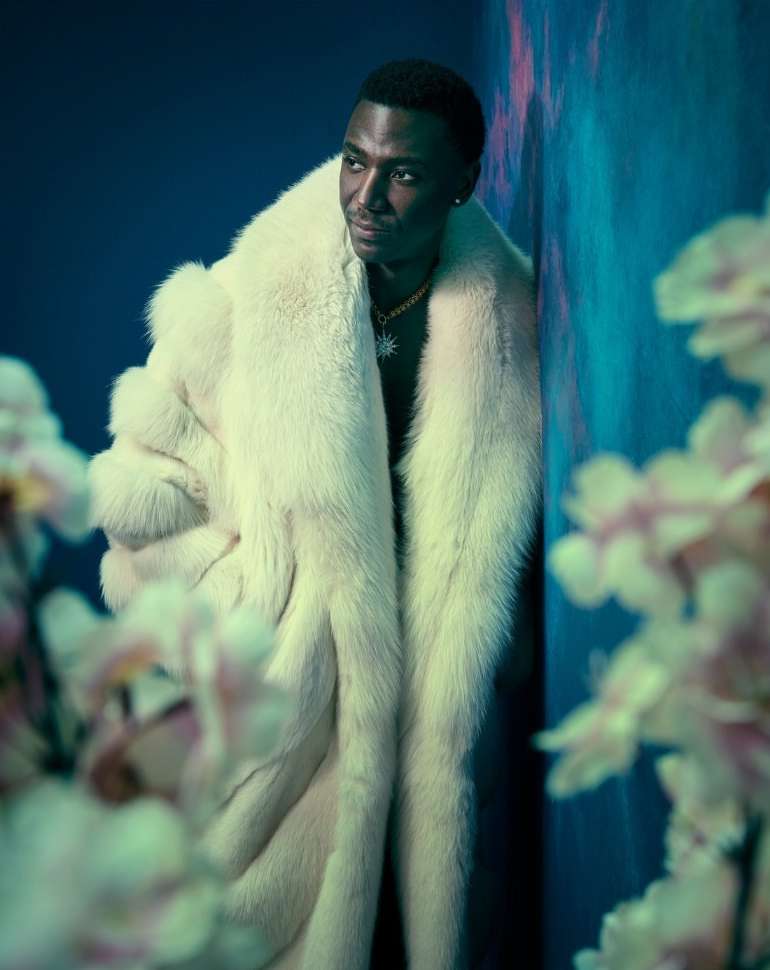Portrait of a Black man wearing a cream-colored fur coat, standing against a blue wall with floral foreground details.