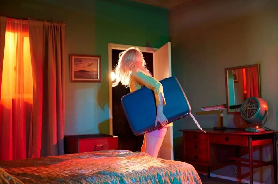 Woman with blonde hair is carrying a blue suitcase in a retro-style bedroom with red curtains, a bed, a desk, and a vintage fan.