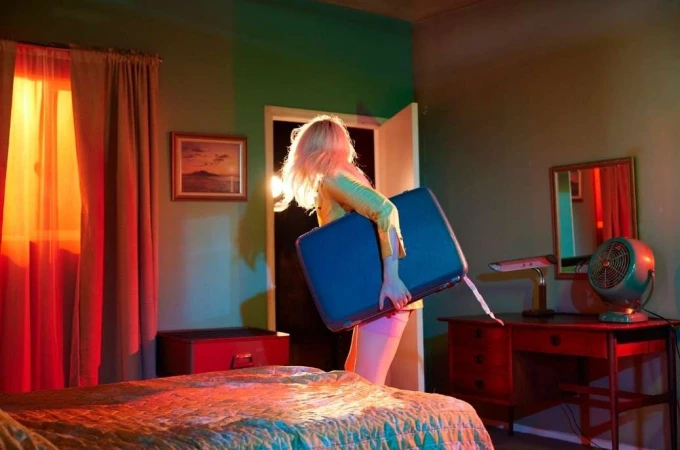 Woman with blonde hair is carrying a blue suitcase in a retro-style bedroom with red curtains, a bed, a desk, and a vintage fan.