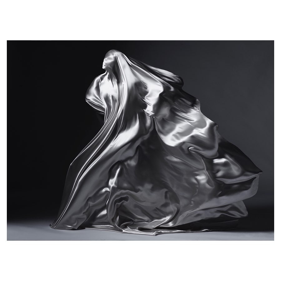 A woman's silhouette is draped in flowing silver fabric against a dark background, creating a dynamic and elegant composition.