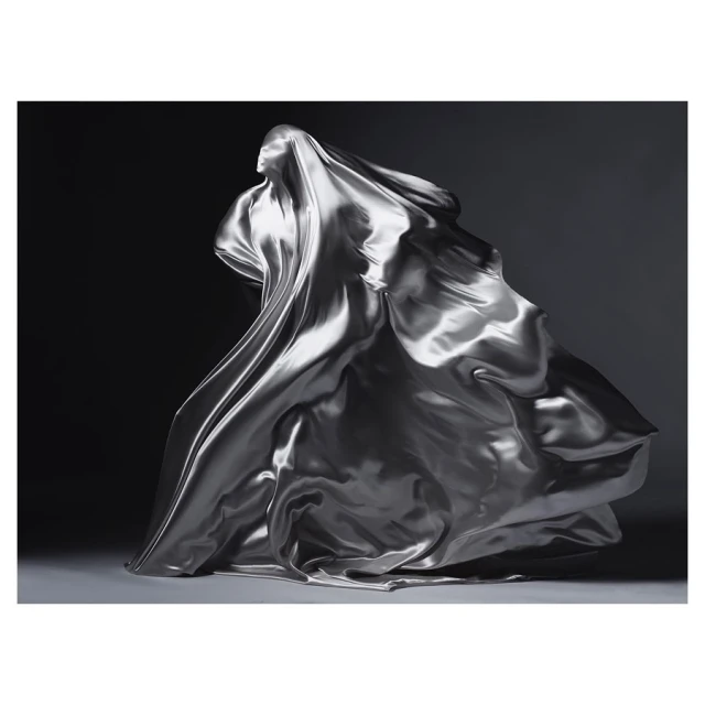 A woman's silhouette is draped in flowing silver fabric against a dark background, creating a dynamic and elegant composition.