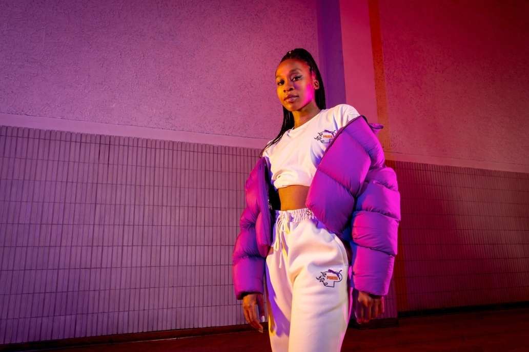 Woman poses in white track suit with violet puffer jacket against textured violet wall in a studio. Fashion shoot with vibrant lighting.