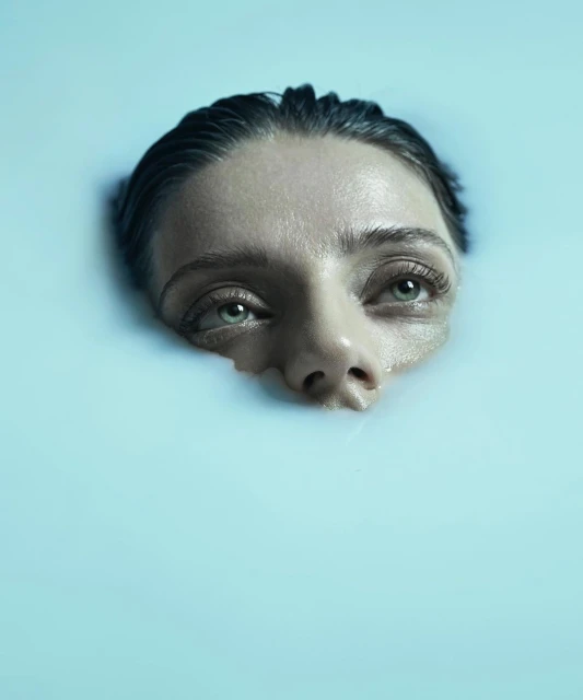 An adult woman is partially submerged in milky blue water, her face and upper chest visible, in a beauty or wellness context.