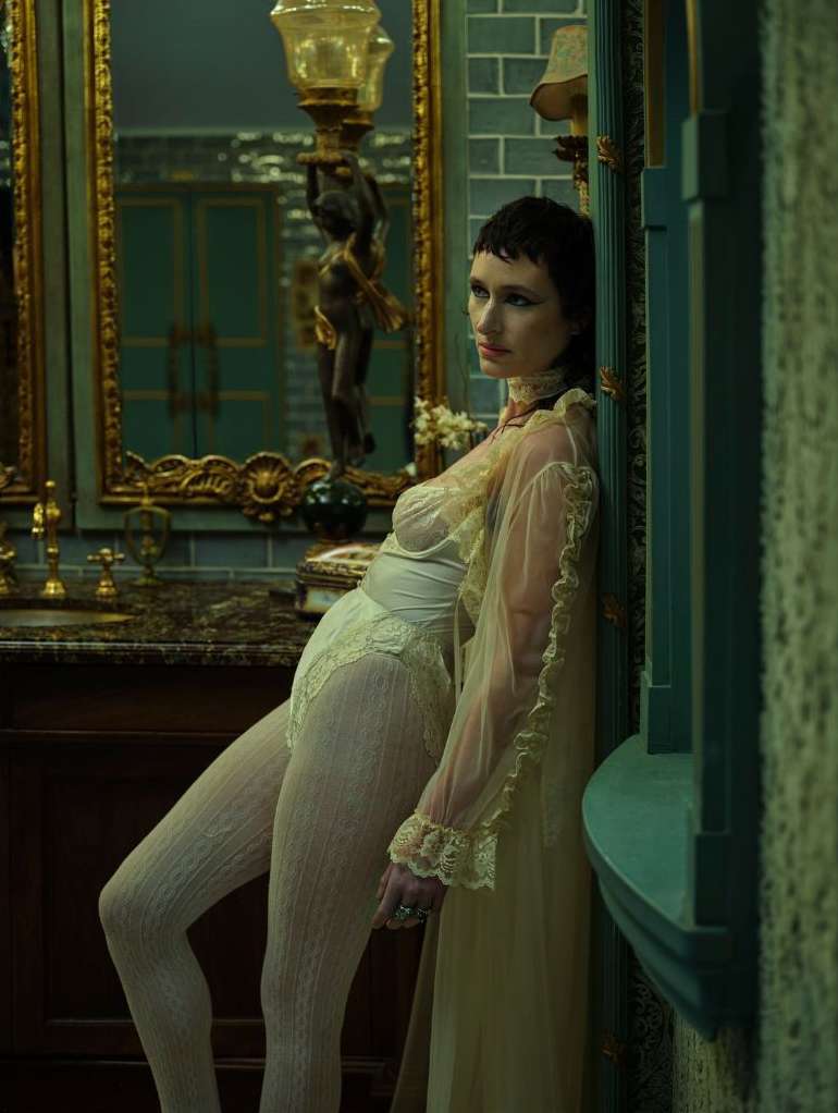 A woman with short, dark hair poses in lingerie and a sheer robe in a vintage-style bathroom with ornate gold and teal details.