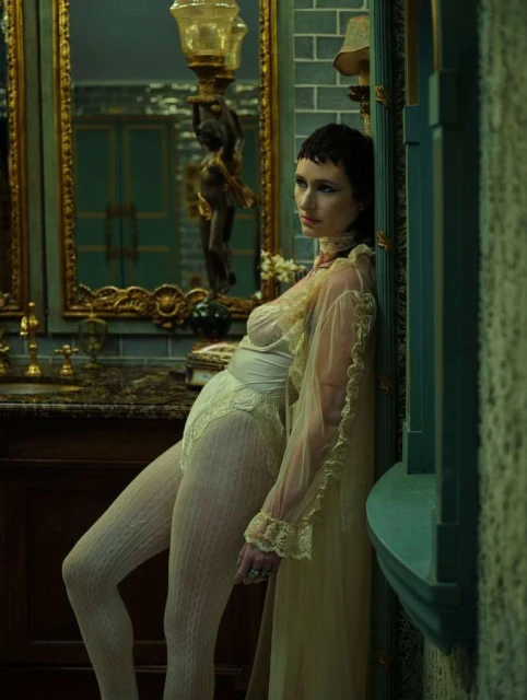 A woman with short, dark hair poses in lingerie and a sheer robe in a vintage-style bathroom with ornate gold and teal details.