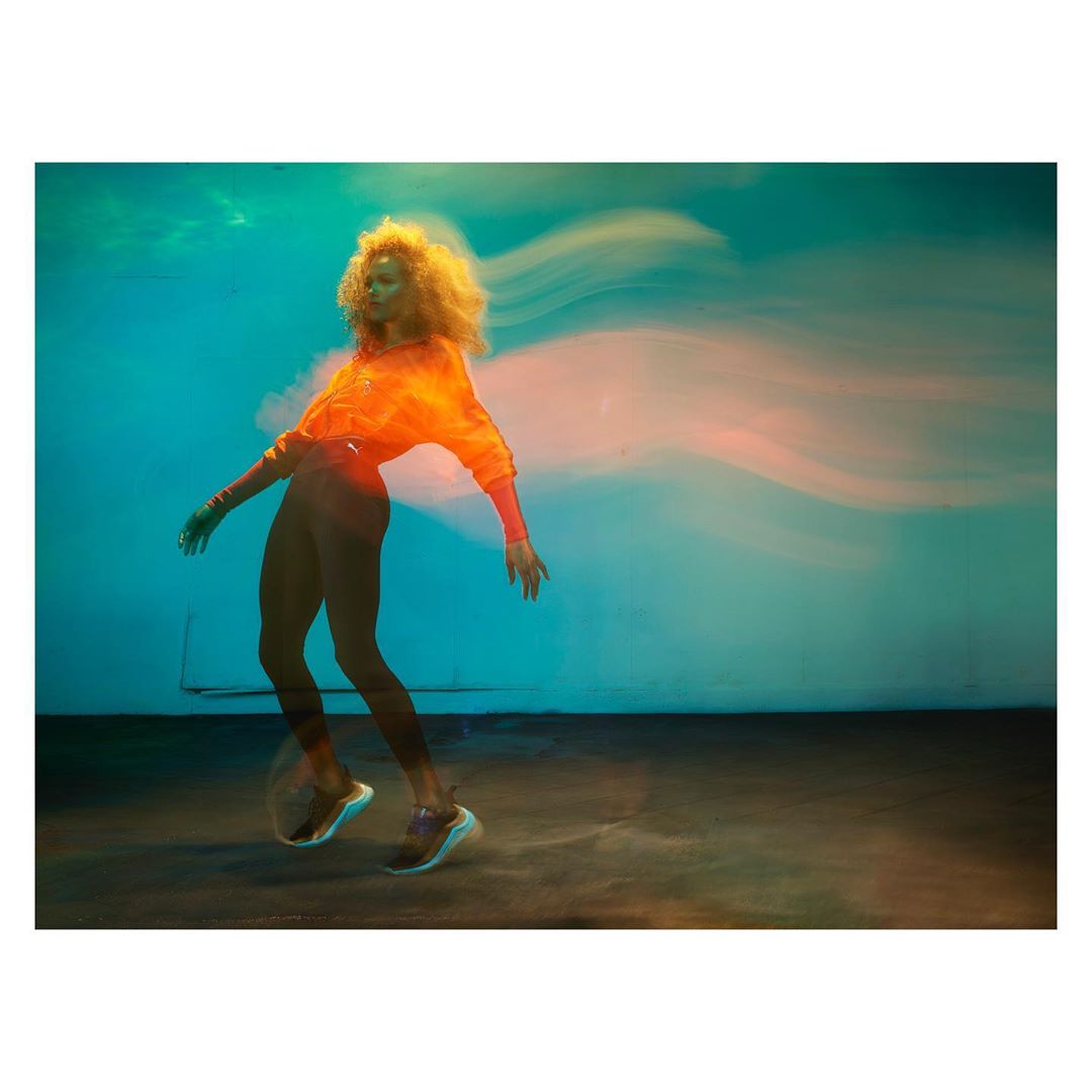 Athletic woman in motion wearing orange top and dark leggings in studio with teal backdrop and motion blur effect showcasing movement.