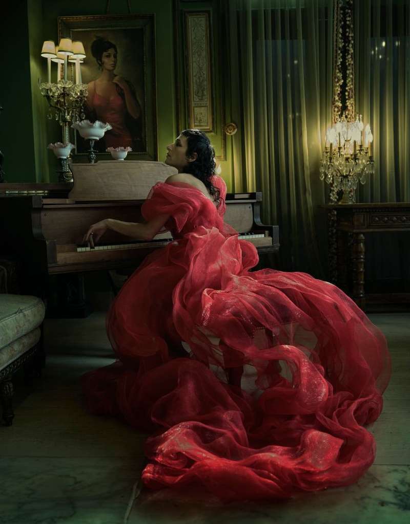 Woman in flowing red gown plays grand piano indoors with framed portrait, ornate decor, soft light, and green walls in background.