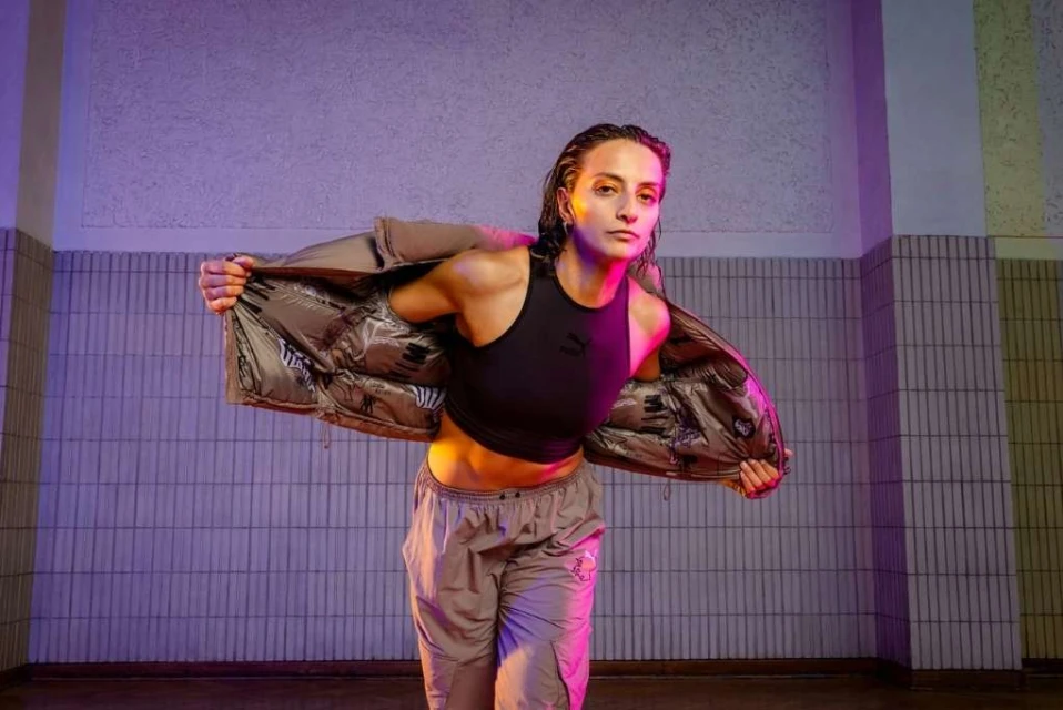 Athletic woman in crop top and track pants poses indoors wearing puffer jacket under purple and yellow lights in a tiled room.