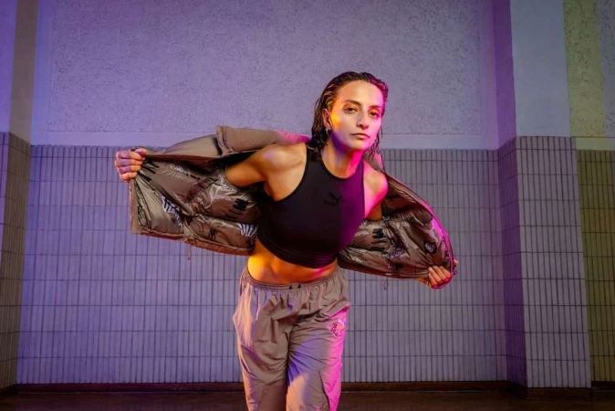 Athletic woman in crop top and track pants poses indoors wearing puffer jacket under purple and yellow lights in a tiled room.