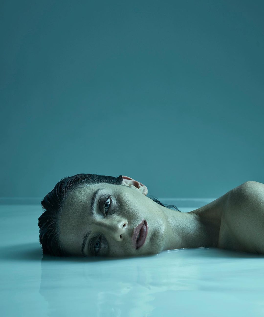 Close-up portrait of a woman submerged in milky water, eyes open, resting her head back, set against a seamless blue background.