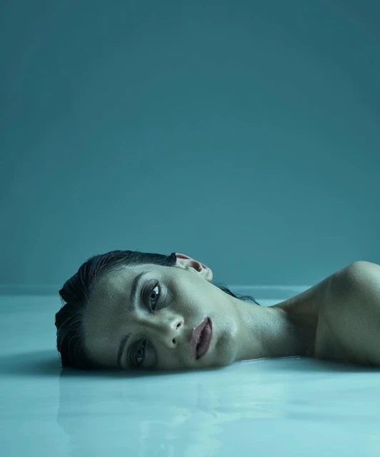 Close-up portrait of a woman submerged in milky water, eyes open, resting her head back, set against a seamless blue background.