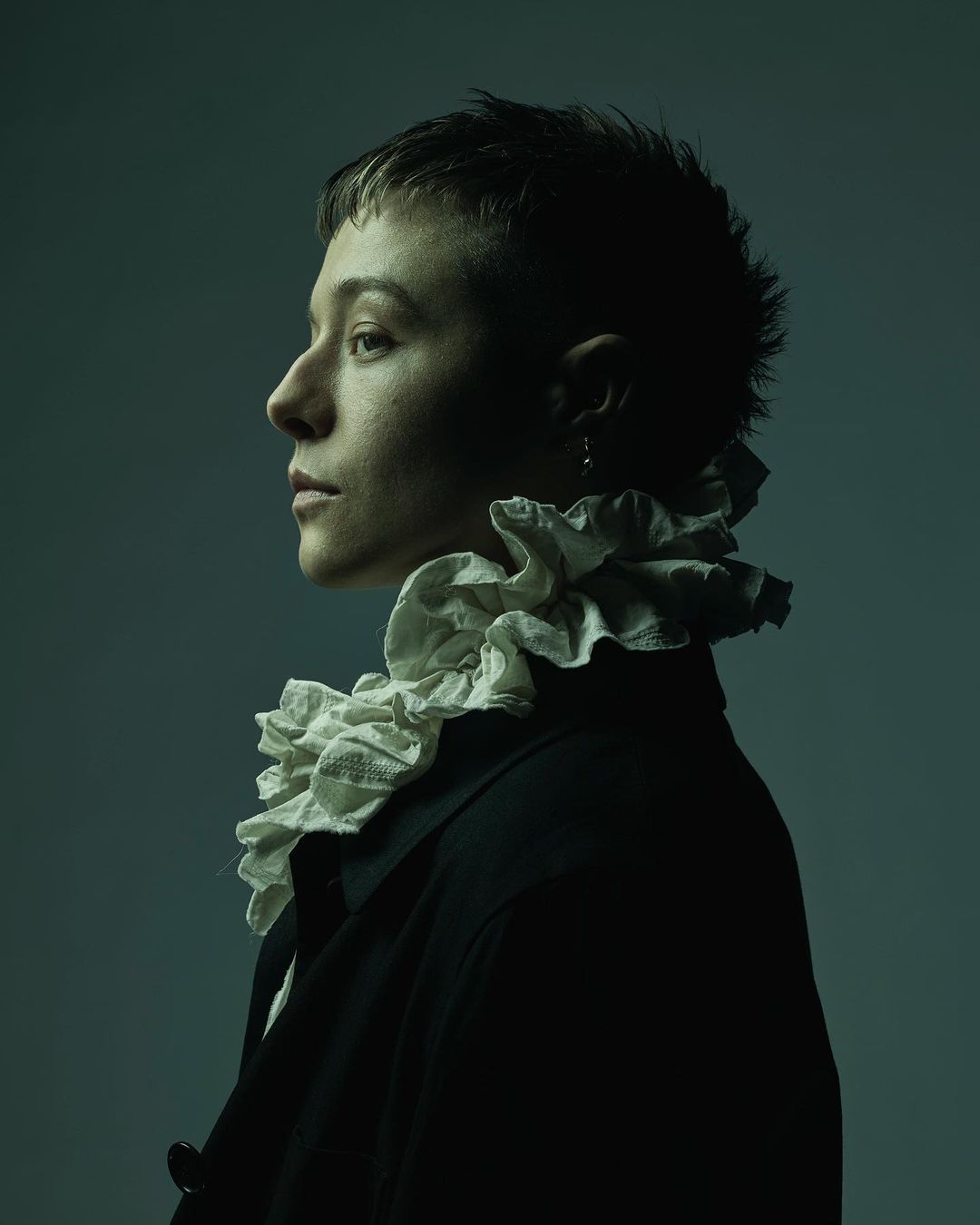 Profile portrait of a person with a short haircut wearing a black coat and white ruffled collar on a muted greenish blue background.