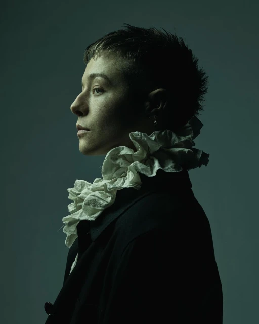 Profile portrait of a person with a short haircut wearing a black coat and white ruffled collar on a muted greenish blue background.