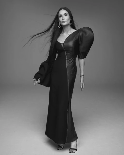 Elegant portrait of adult woman with long black hair, wearing a full-length black gown with one long sleeve, posing in a studio setting.