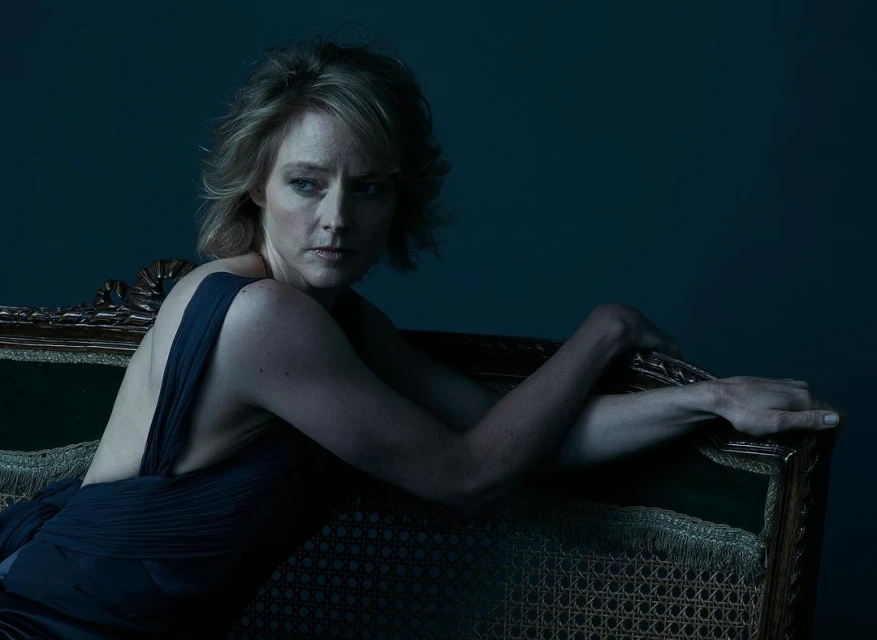 Portrait of a woman with short blonde hair, wearing a dark dress, reclining on an ornate couch against a dark green background.