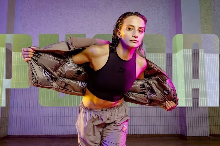 Athletic woman poses in a black crop top and grey sweatpants, partially wearing a jacket in a brightly lit indoor setting.