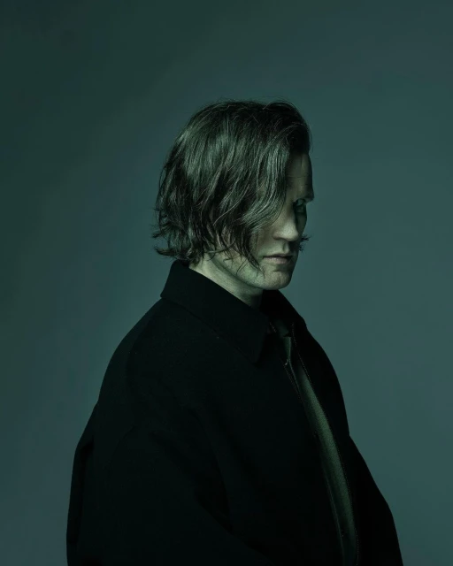 Profile portrait of an adult male with shoulder-length dark hair wearing a black coat against a green gradient background in studio lighting.