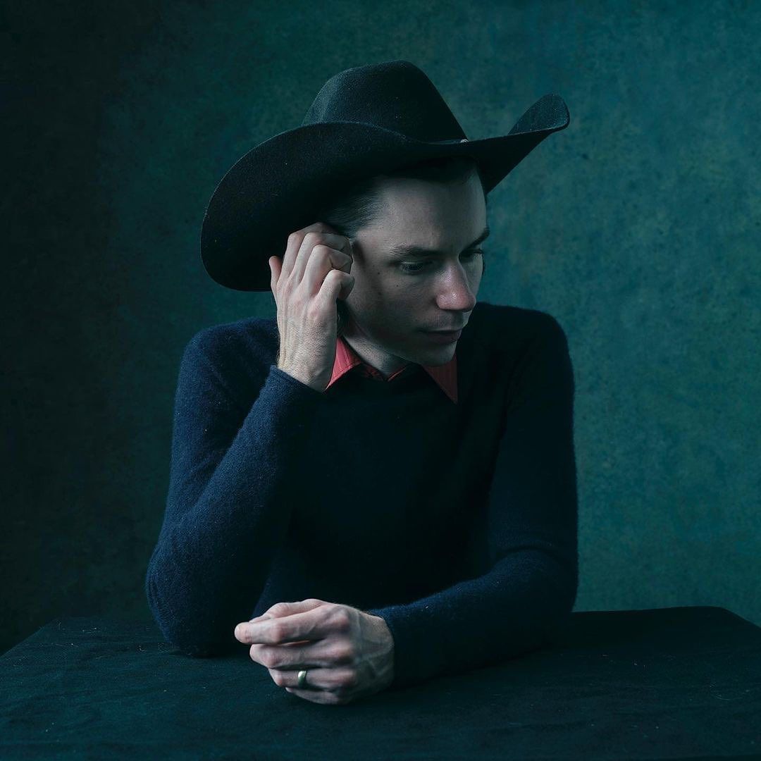 Indoor portrait of an adult male wearing a black cowboy hat, dark sweater, and red shirt, posing at a table against a textured backdrop.