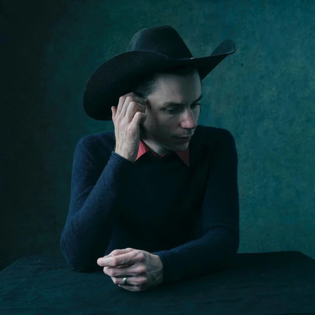 Indoor portrait of an adult male wearing a black cowboy hat, dark sweater, and red shirt, posing at a table against a textured backdrop.