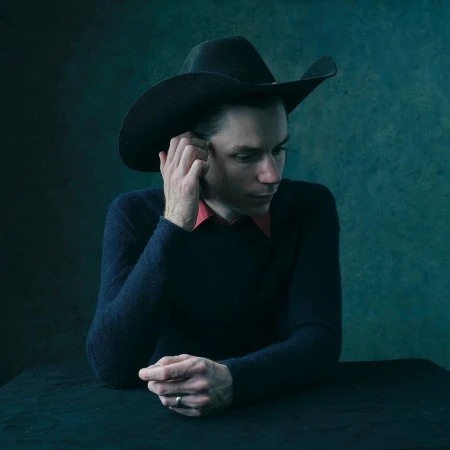 Indoor portrait of an adult male wearing a black cowboy hat, dark sweater, and red shirt, posing at a table against a textured backdrop.