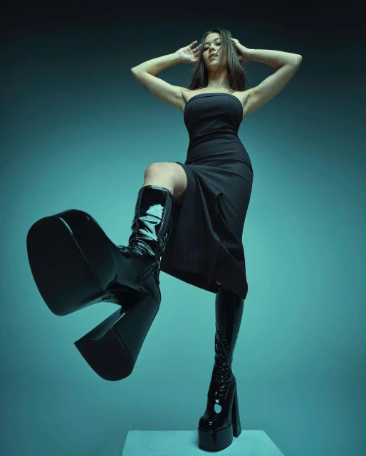 Low-angle studio shot of a woman in a black dress and shiny, oversized black platform boots striking a dynamic pose against a gradient teal background.
