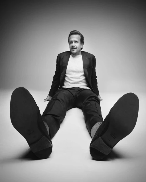 Black and white shot of a seated adult male wearing a blazer and trousers, with the camera focusing on his outstretched shoes.
