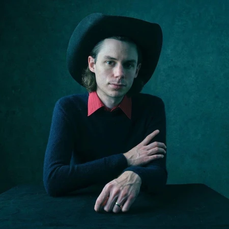 Portrait of a man in a black cowboy hat, navy sweater, and red collared shirt against a dark textured teal background.