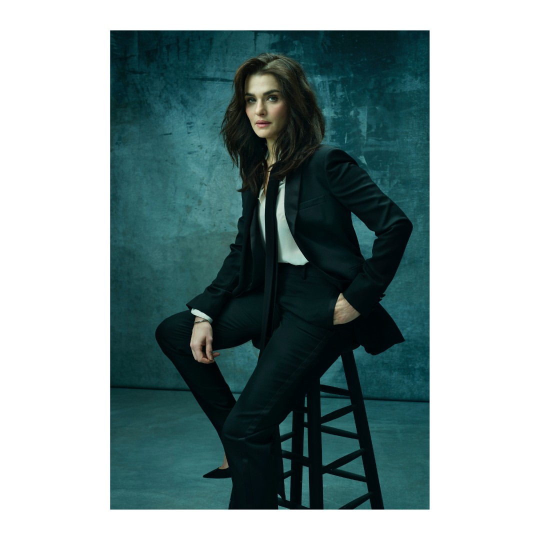 Portrait of Rachel Weisz seated in a dark suit, tie, and white shirt against a mottled blue-grey backdrop, striking a poised, professional pose.